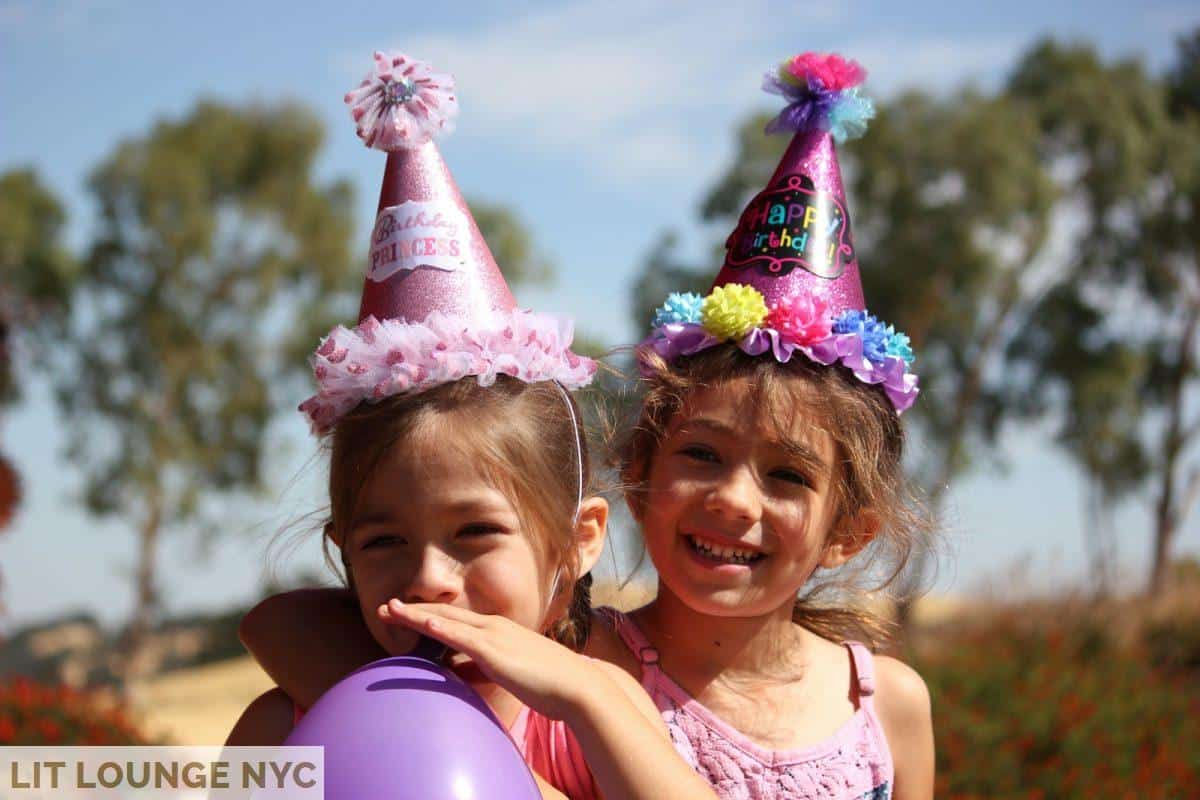 girl in left blowing purple balloon beside girl wearing pink birthday hat